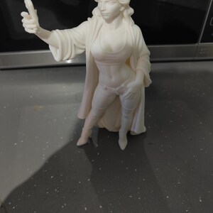 K&ouml;nnte beinhalten: Eine wei&szlig;e Figur der Freiheitsstatue, mit einer Krone aus Stacheln und flie&szlig;enden Gew&auml;ndern. Die Figur ist in einem modernen, stilisierten Design gehalten, mit erhobener Hand. Die Figur steht auf einer dunklen Oberfl&auml;che.
