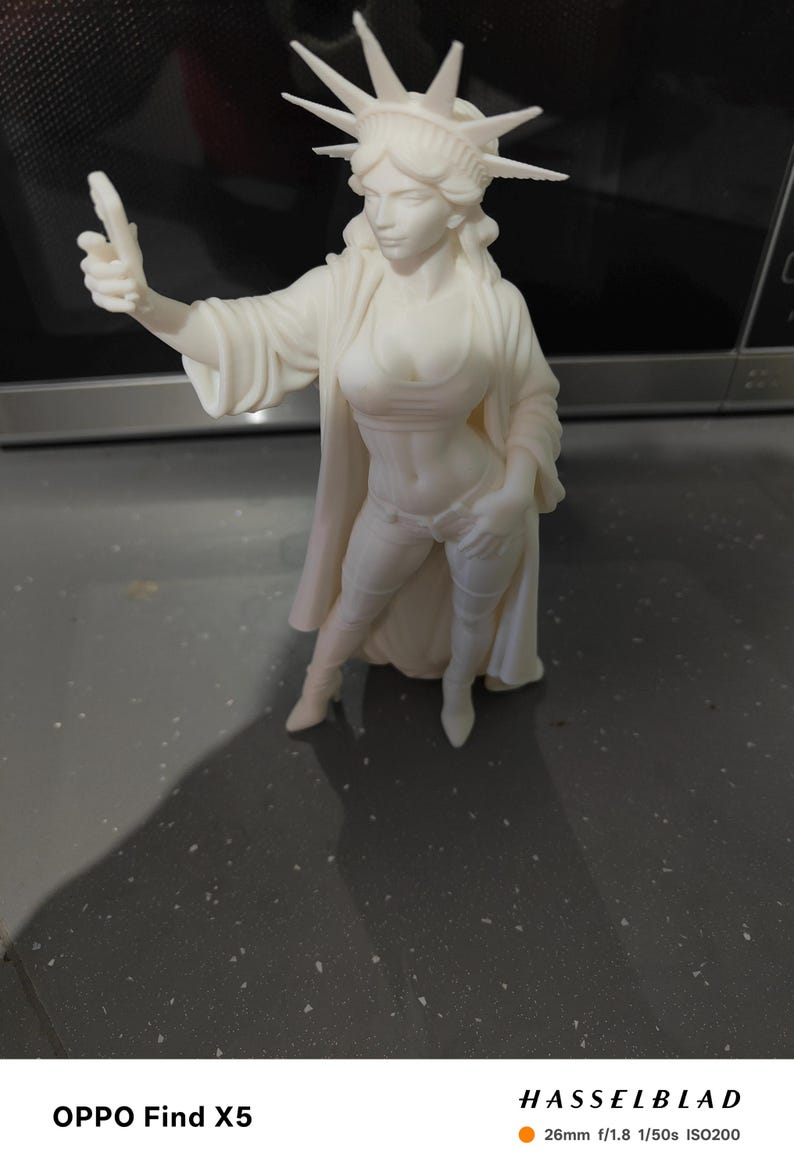 K&ouml;nnte beinhalten: Eine wei&szlig;e Figur der Freiheitsstatue, mit einer Krone aus Stacheln und flie&szlig;enden Gew&auml;ndern. Die Figur ist in einem modernen, stilisierten Design gehalten, mit erhobener Hand. Die Figur steht auf einer dunklen Oberfl&auml;che.