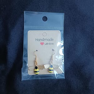 May include: Bee-shaped earrings with gold hooks and wings. The bees have black and yellow striped bodies, presented in a clear plastic bag with a card reading "Handmade with love".