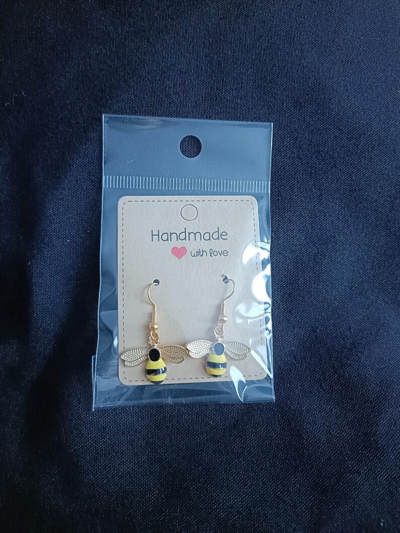 May include: Bee-shaped earrings with gold hooks and wings. The bees have black and yellow striped bodies, presented in a clear plastic bag with a card reading "Handmade with love".