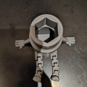 May include: A gray, 3D-printed socket wrench figure with articulated legs and arms. The wrench head is a hexagonal shape. The figure is standing on a dark surface.