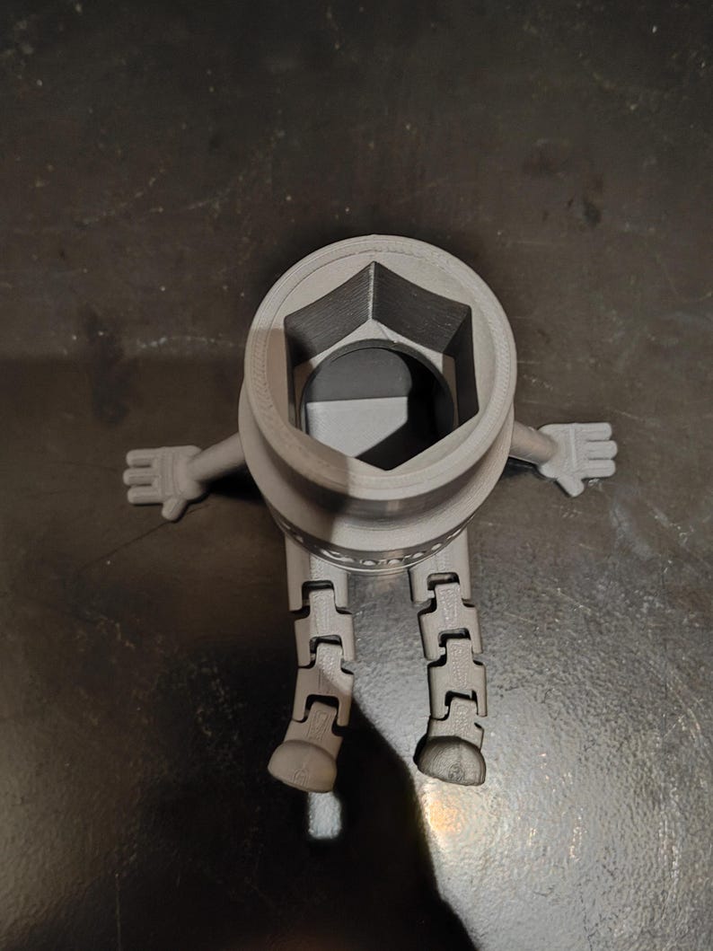 May include: A gray, 3D-printed socket wrench figure with articulated legs and arms. The wrench head is a hexagonal shape. The figure is standing on a dark surface.