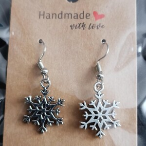 May include: Silver snowflake earrings hanging from silver hooks. The earrings are displayed on a brown card with the words "Handmade with love" printed on it. The earrings are a winter-themed accessory.