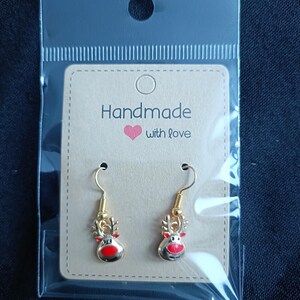 May include: A pair of gold-colored reindeer earrings with red noses and antlers, displayed on a tan card that reads "Handmade with love." The earrings are packaged in a clear plastic bag.