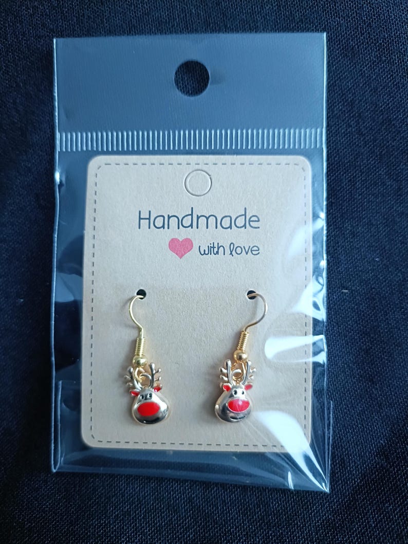 May include: A pair of gold-colored reindeer earrings with red noses and antlers, displayed on a tan card that reads "Handmade with love." The earrings are packaged in a clear plastic bag.