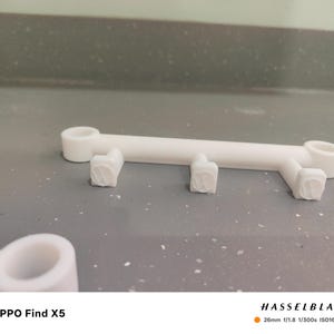 May include: A white, 3D-printed object with a linear design. It features a central bar with two circular ends and three square, textured supports. The object is set against a gray background. The text "OPPO Find X5" is visible.