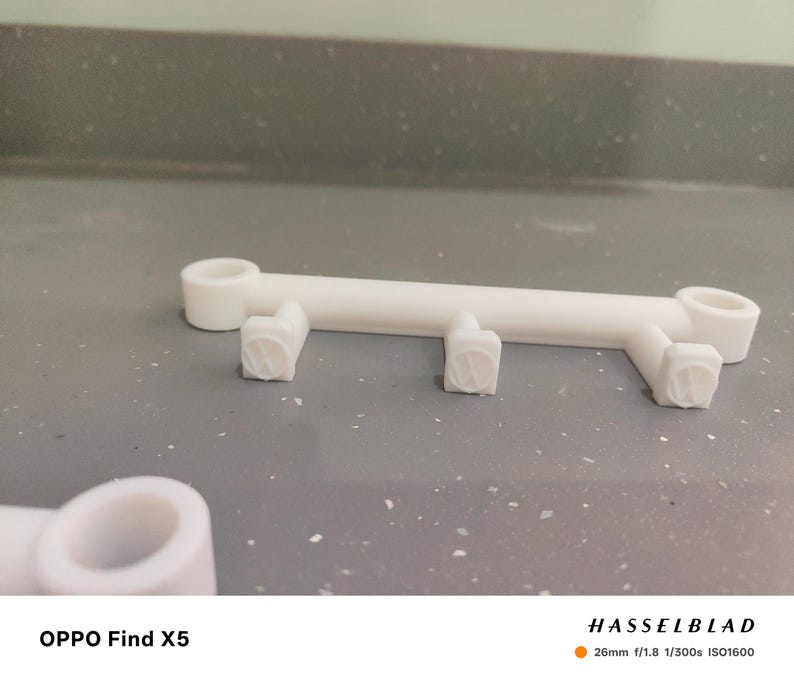 May include: A white, 3D-printed object with a linear design. It features a central bar with two circular ends and three square, textured supports. The object is set against a gray background. The text "OPPO Find X5" is visible.