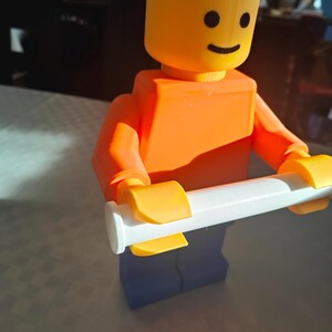 May include: A 3D-printed Lego figure in orange, yellow, and blue, holding a white object. The figure has a smiling face. The image is well-lit, with a focus on the toy. The background is blurred.