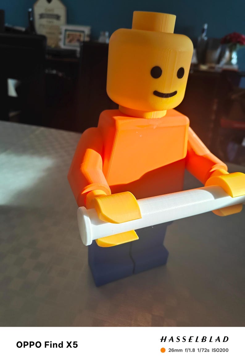 May include: A 3D-printed Lego figure in orange, yellow, and blue, holding a white object. The figure has a smiling face. The image is well-lit, with a focus on the toy. The background is blurred.