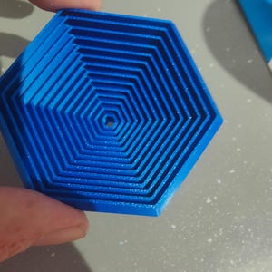 May include: A blue, hexagonal object with a layered design. The object has a series of concentric, hexagonal shapes, creating a 3D effect. The object is held by a person's hand. The image is taken with an OPPO Find X5 camera.