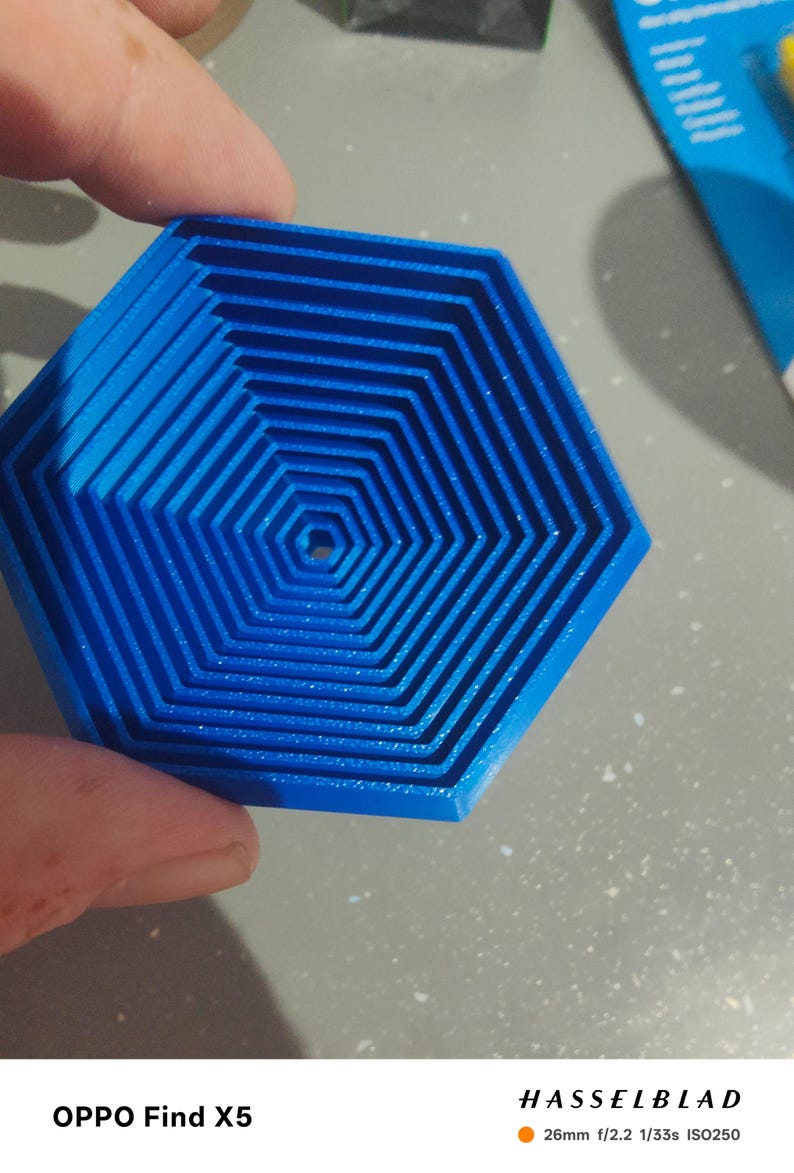 May include: A blue, hexagonal object with a layered design. The object has a series of concentric, hexagonal shapes, creating a 3D effect. The object is held by a person's hand. The image is taken with an OPPO Find X5 camera.