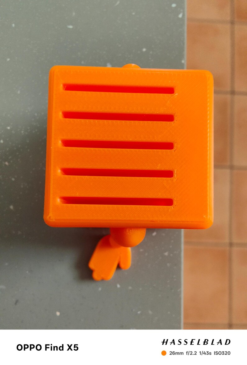 May include: Bright orange, square-shaped object with horizontal slots, and a small, rounded appendage. The object is resting on a gray surface. The image also includes the text "OPPO Find X5" and "HASSELBLAD".