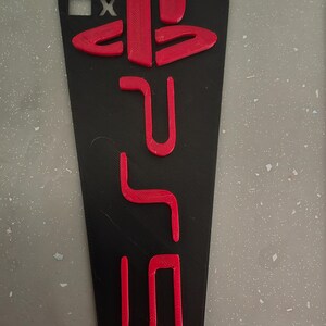 May include: Black rectangular phone case with red PlayStation 5 logo and symbols. The case has a matte finish and is designed to fit a smartphone. The design includes the PlayStation symbols and the PS5 logo.