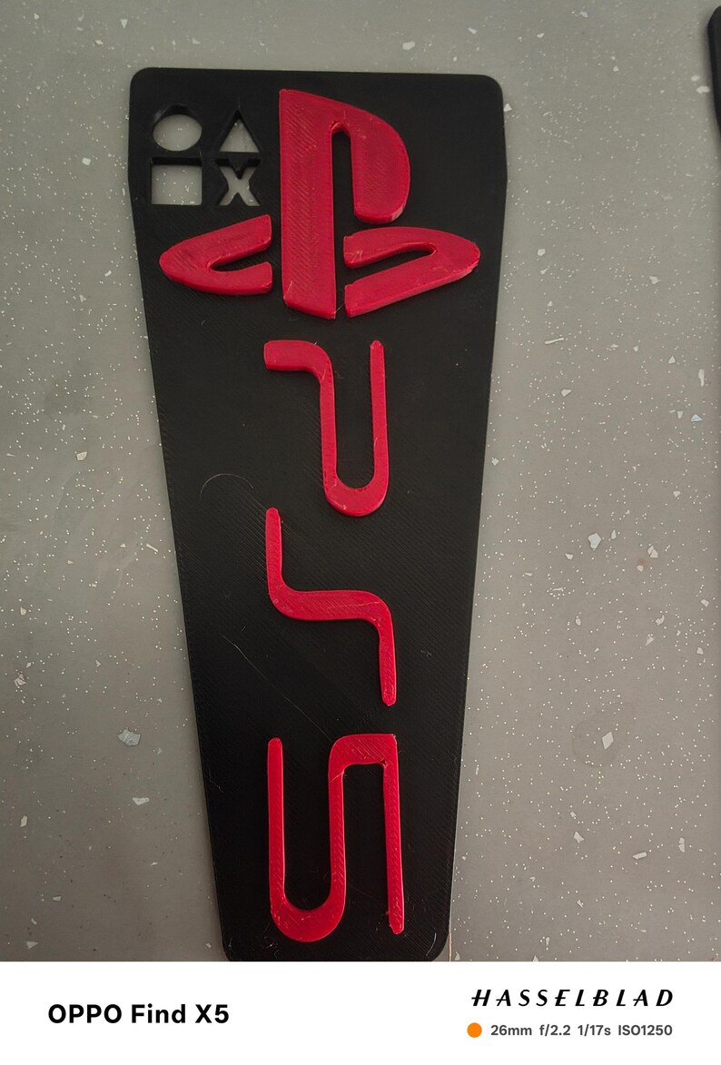 May include: Black rectangular phone case with red PlayStation 5 logo and symbols. The case has a matte finish and is designed to fit a smartphone. The design includes the PlayStation symbols and the PS5 logo.