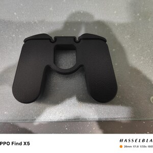 K&ouml;nnte beinhalten: Ein mattschwarzes Telefonzubeh&ouml;r in Form eines Gamecontrollers liegt auf einer hellgrauen Oberfl&auml;che. Das Zubeh&ouml;r hat eine strukturierte Oberfl&auml;che und einen mittigen Ausschnitt. Der Text "OPPO Find X5" ist unten links sichtbar.