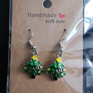 Op de afbeelding: Een paar kerstboom oorbellen met groene emaille, rode en gele accenten en zilverkleurige haken. De oorbellen worden getoond op een bruine kaart met de tekst "Handmade with love."
