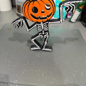 May include: A Halloween-themed decoration featuring a pumpkin head atop a skeleton body. The pumpkin is orange with a black stem and facial features. The skeleton is black and white, with arms outstretched, pointing. A fun, festive item.
