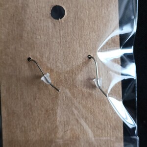 May include: A pair of silver-toned earrings with clear plastic ear protectors, displayed on a brown cardboard backing inside a clear plastic bag. The earrings have a simple hook design.