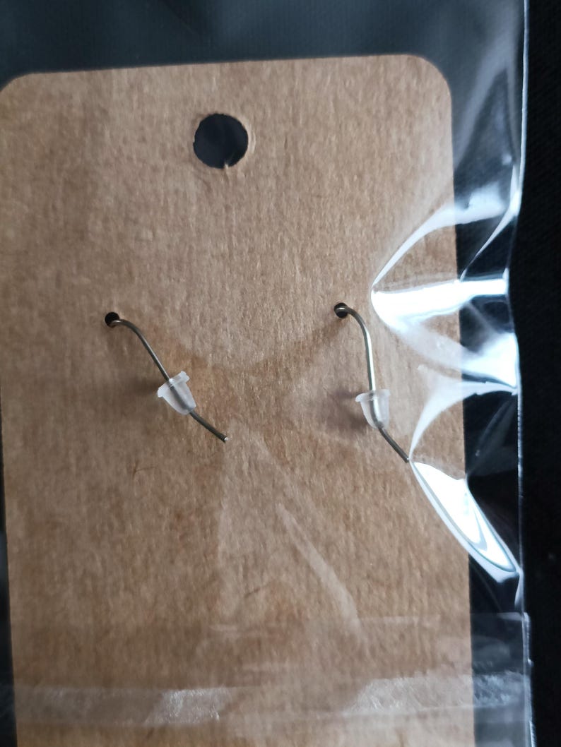 May include: A pair of silver-toned earrings with clear plastic ear protectors, displayed on a brown cardboard backing inside a clear plastic bag. The earrings have a simple hook design.