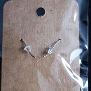 May include: A pair of silver earrings with clear, rounded stoppers, presented on a brown cardboard earring card. The card features a small hole at the top and two smaller holes for the earring hooks.