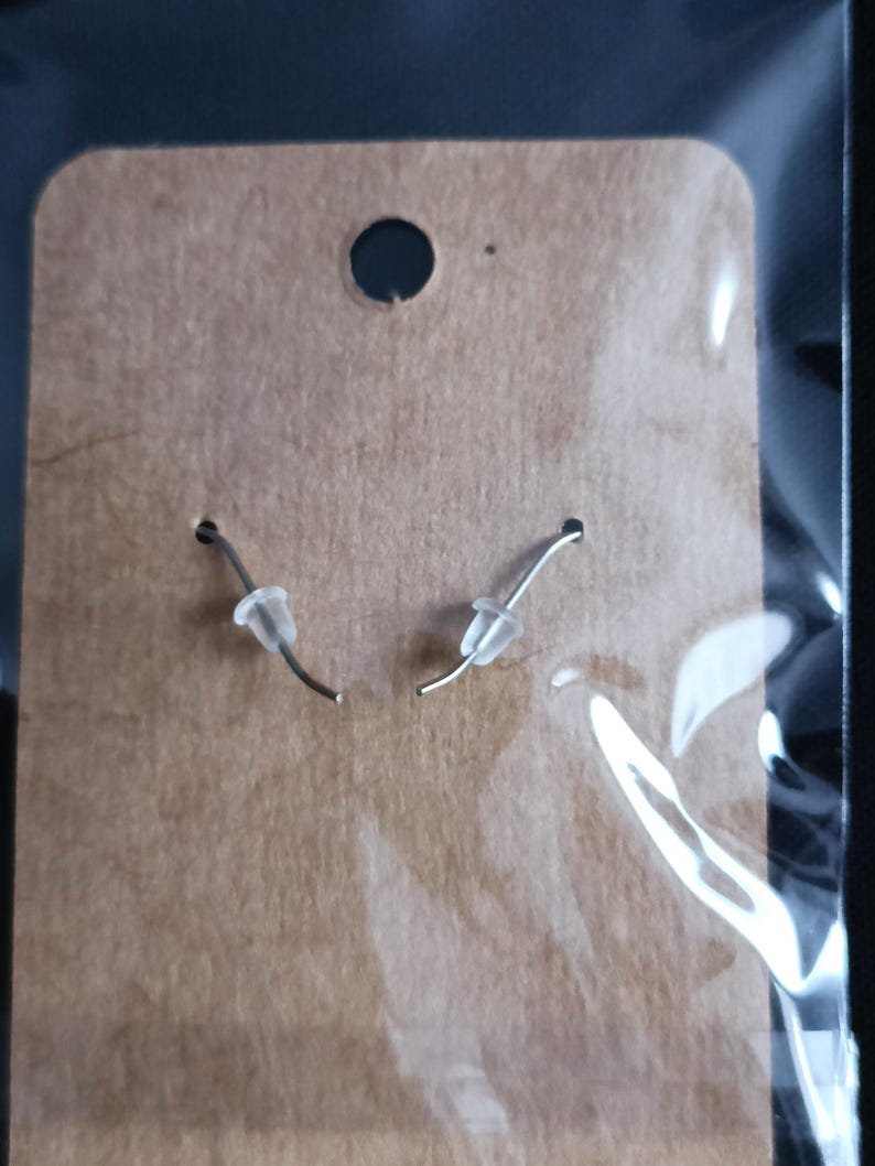 May include: A pair of silver earrings with clear, rounded stoppers, presented on a brown cardboard earring card. The card features a small hole at the top and two smaller holes for the earring hooks.