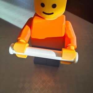 May include: A 3D-printed Lego figure in bright orange and yellow, holding a white cylinder. The figure has a smiling face and is standing on a dark base. The image is well-lit, with a focus on the toy.