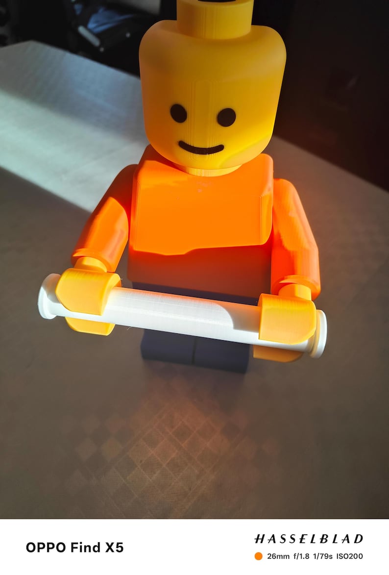 May include: A 3D-printed Lego figure in bright orange and yellow, holding a white cylinder. The figure has a smiling face and is standing on a dark base. The image is well-lit, with a focus on the toy.