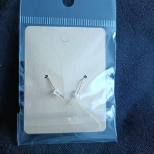 May include: A pair of gold-colored earrings with clear plastic stoppers are displayed on a tan earring card inside a clear plastic bag. The bag has a hole at the top for hanging. The background is a dark blue fabric.