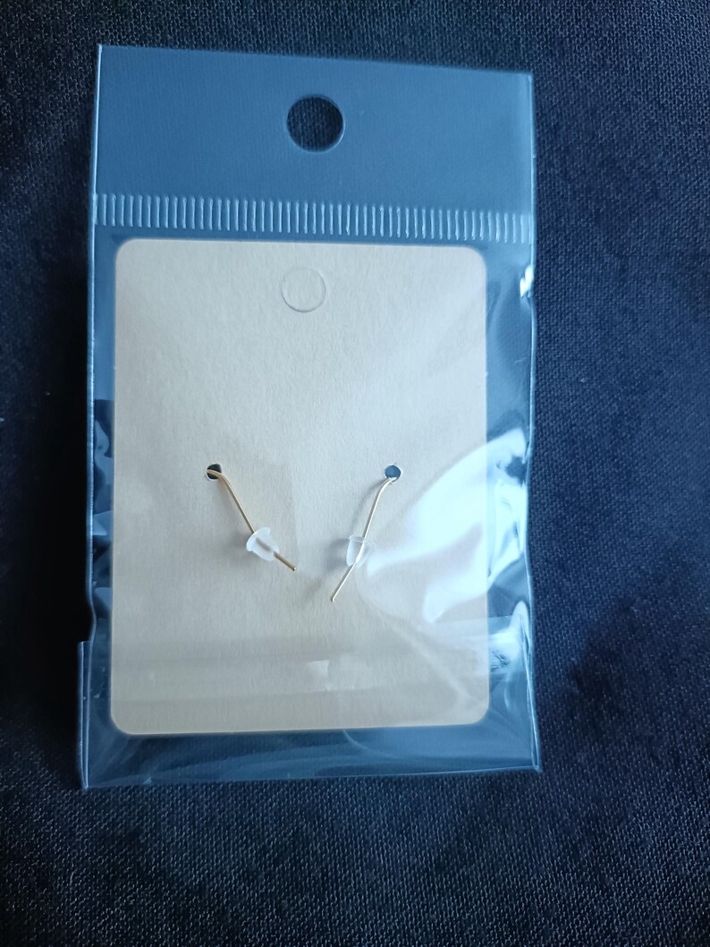 May include: A pair of gold-colored earrings with clear plastic stoppers are displayed on a tan earring card inside a clear plastic bag. The bag has a hole at the top for hanging. The background is a dark blue fabric.