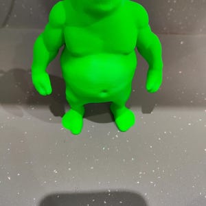 K&ouml;nnte beinhalten: Eine hellgr&uuml;ne Figur einer Figur mit gro&szlig;en Ohren, einem breiten Mund und einem muskul&ouml;sen K&ouml;rperbau. Die Figur steht auf einer grauen Oberfl&auml;che. Die Figur ist eine Zeichentrickfigur.