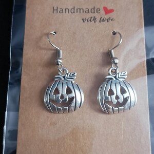 May include: Silver-coloured pumpkin earrings with jack-o'-lantern faces. The earrings hang from silver hooks and are displayed on a brown card with the text "Handmade with love".