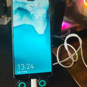 K&ouml;nnte beinhalten: Ein Smartphone mit einem hellblauen Bildschirm, der die Uhrzeit 13:24 und das Datum, Sonntag, 14. September, anzeigt. Das Telefon befindet sich in einem schwarzen St&auml;nder mit gr&uuml;nen Akzenten und einem wei&szlig;en Ladekabel. Das Telefonmodell ist OPPO Find X5.
