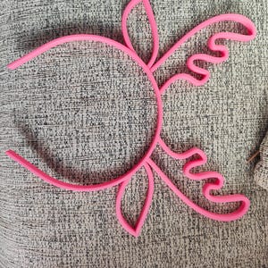 Puede incluir: Una diadema rosa brillante con un dise&ntilde;o floral. La diadema tiene una base circular con formas curvas en forma de p&eacute;talo que se extienden hacia afuera. La imagen fue tomada con una c&aacute;mara OPPO Find X5.
