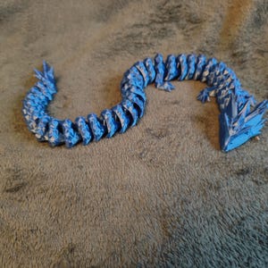 May include: A blue and white articulated dragon toy. The dragon is made of many small, connected pieces, allowing it to be posed. The dragon's head and tail have a spiky design. The toy is lying on a gray, textured surface.