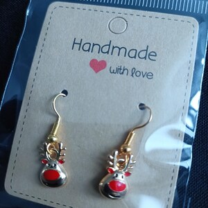 May include: A pair of gold-colored reindeer earrings with red noses and antlers, displayed on a brown card that reads "Handmade with love". The earrings are in a clear plastic package.
