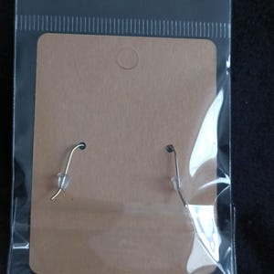 May include: Earrings displayed on a brown cardboard backing, enclosed in a clear plastic bag. The earrings have silver-colored hooks. The packaging has a small hole at the top for hanging.