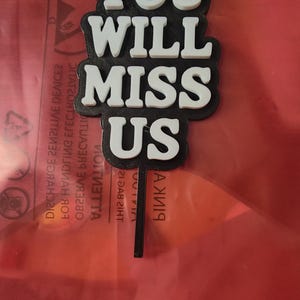 May include: A black and white cake topper with the words "YOU WILL MISS US". The text is in a bold, sans-serif font. The topper is on a black stick, and the background is red.
