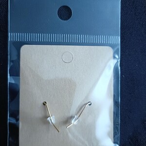 May include: A pair of gold-colored fish hook earrings with clear plastic stoppers, displayed on a tan card inside a clear plastic bag. The earrings are simple and elegant, suitable for everyday wear.
