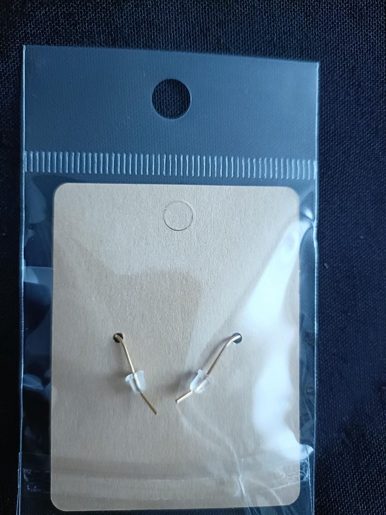 May include: A pair of gold-colored fish hook earrings with clear plastic stoppers, displayed on a tan card inside a clear plastic bag. The earrings are simple and elegant, suitable for everyday wear.