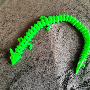 May include: A bright green, articulated dragon toy. The dragon is made of plastic and has a segmented body, allowing for flexible movement. The dragon is lying on a brown, textured surface.