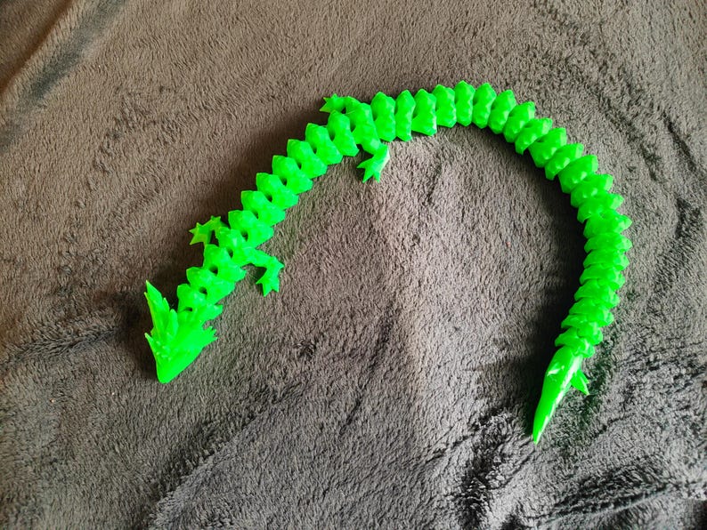 May include: A bright green, articulated dragon toy. The dragon is made of plastic and has a segmented body, allowing for flexible movement. The dragon is lying on a brown, textured surface.