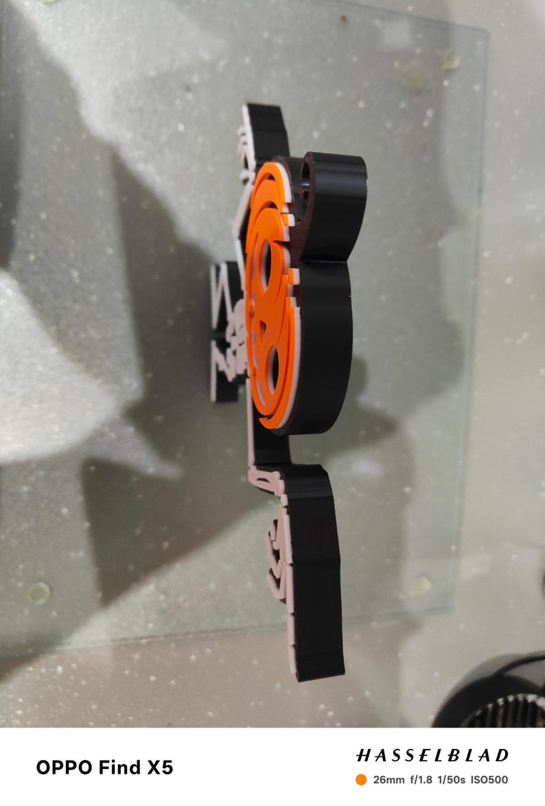 May include: A black, white, and orange film reel logo. The logo is made of layered materials and is mounted on a clear surface. The logo is a stylized representation of a film reel, with the word "film" incorporated into the design.