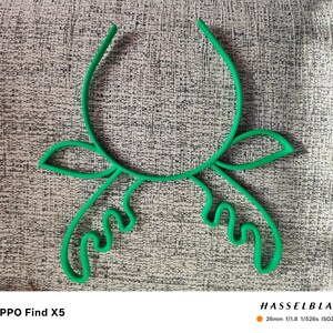 Puede incluir: Un objeto verde brillante con forma floral abstracta. El dise&ntilde;o presenta l&iacute;neas curvas y formas de hojas, creando una forma bot&aacute;nica estilizada. El objeto se encuentra sobre un fondo gris texturizado. La imagen tambi&eacute;n incluye especificaciones de la c&aacute;mara.