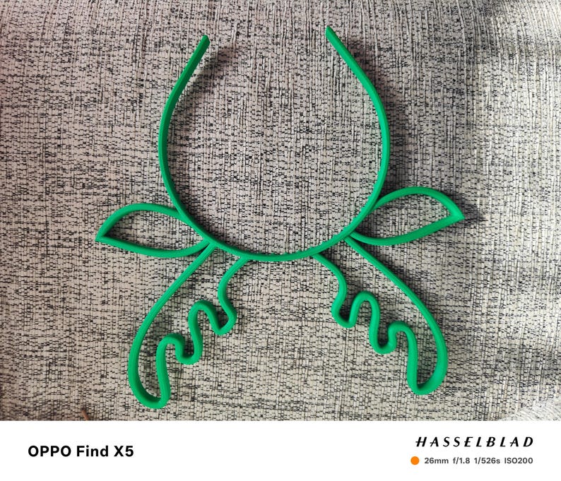 Puede incluir: Un objeto verde brillante con forma floral abstracta. El dise&ntilde;o presenta l&iacute;neas curvas y formas de hojas, creando una forma bot&aacute;nica estilizada. El objeto se encuentra sobre un fondo gris texturizado. La imagen tambi&eacute;n incluye especificaciones de la c&aacute;mara.