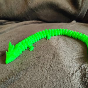 May include: A bright green, articulated dragon toy, 3D-printed with a segmented body. The dragon is posed on a brown, textured surface. The dragon has a detailed head and tail.