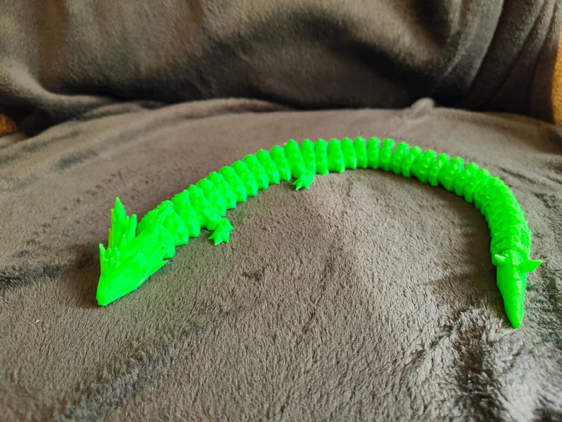 May include: A bright green, articulated dragon toy, 3D-printed with a segmented body. The dragon is posed on a brown, textured surface. The dragon has a detailed head and tail.