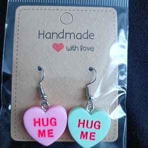 May include: A pair of heart-shaped earrings in a clear plastic package. One earring is pink with "HUG ME" in red, the other is mint green with the same text. The earrings hang from silver-colored hooks. The packaging includes a brown tag that reads "Handmade with love."