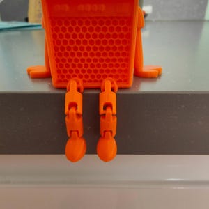 May include: An orange, 3D-printed object resembling a small robot or character. The body is a rectangular shape with a honeycomb pattern, and it has jointed legs hanging down. The object is sitting on a gray surface.