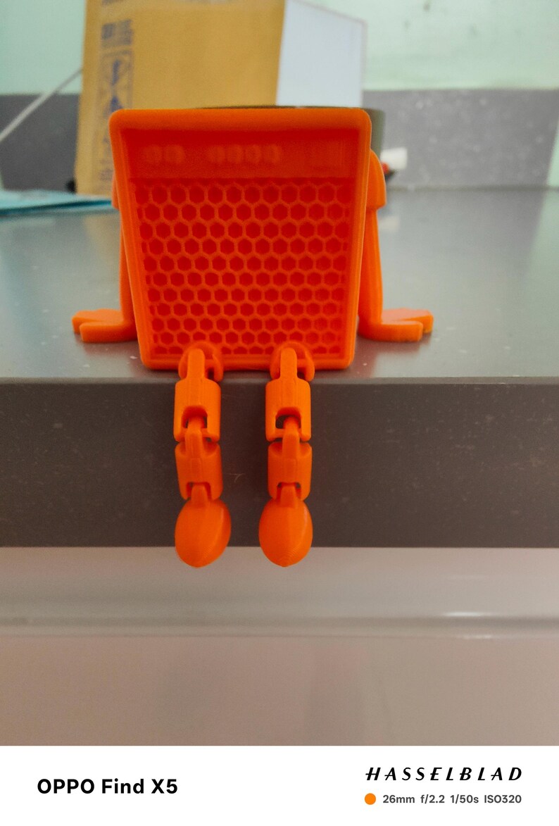 May include: An orange, 3D-printed object resembling a small robot or character. The body is a rectangular shape with a honeycomb pattern, and it has jointed legs hanging down. The object is sitting on a gray surface.