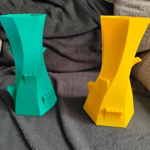 May include: Two colorful, abstract vases. One is teal, the other is yellow. Both have a twisted design with small, rectangular protrusions. The vases are made of a solid material and sit on a gray surface.